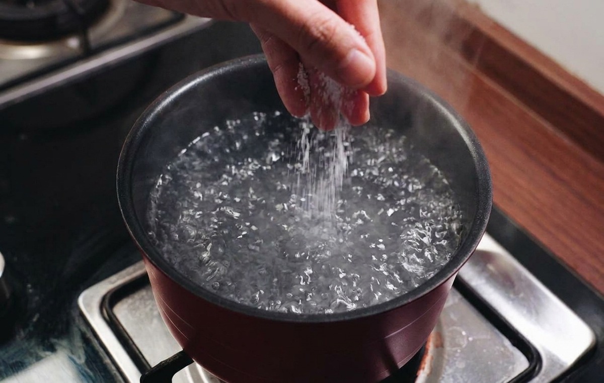 Une main saupoudrant du sel dans une grande casserole deau bouillante sur une cuisinière.