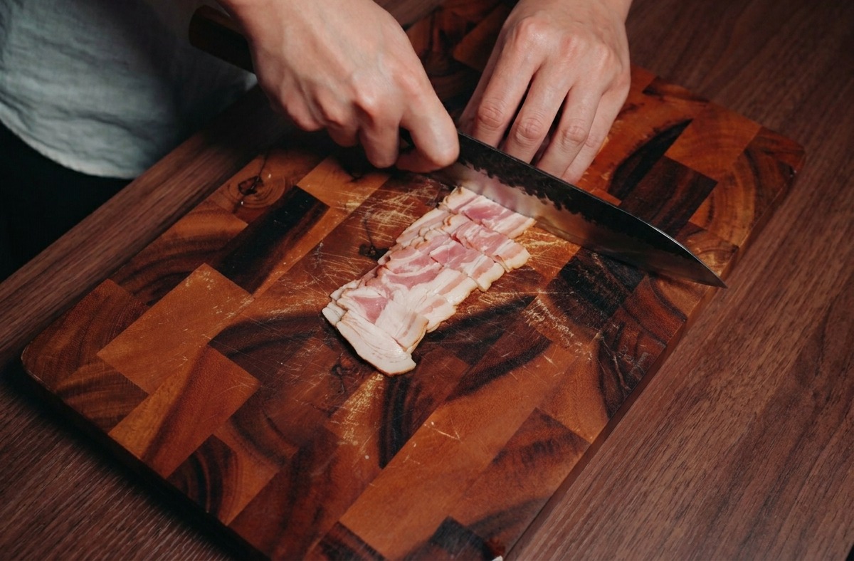Une personne utilisant un couteau de chef pour couper des tranches de bacon en petits carrés sur une planche à découper.