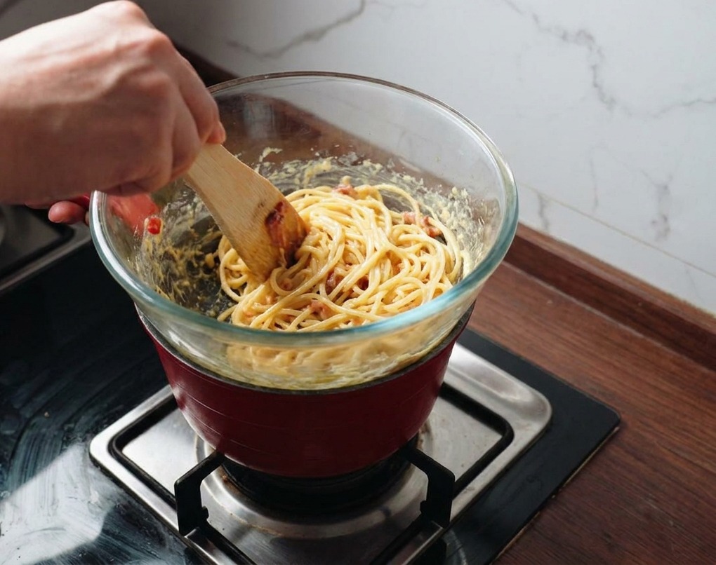 Une personne utilisant une spatule pour mélanger les pâtes et le bacon dans une sauce dorée au-dessus de la vapeur.