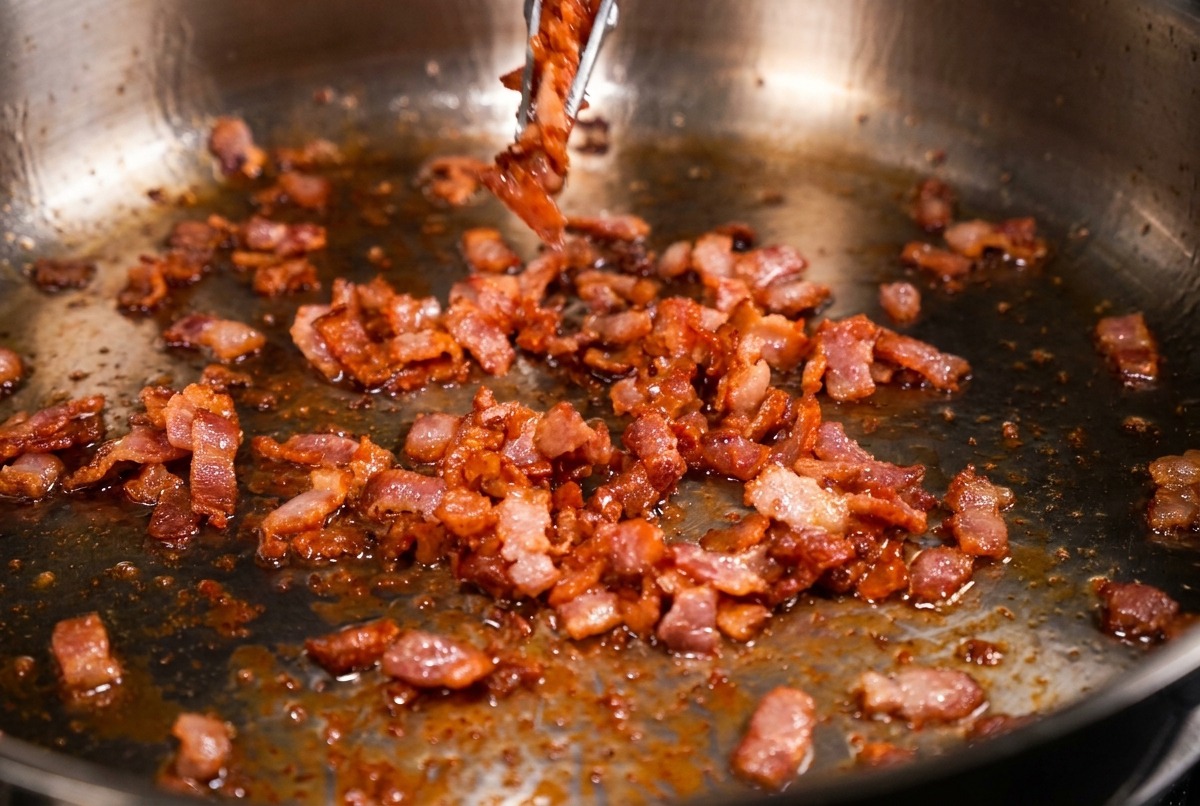 Poêle en inox remplie de petits morceaux de bacon bien dorés, certains étant retirés avec une pince en métal.