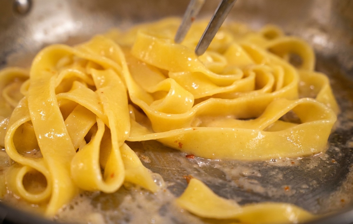 Pince de cuisine remuant des pâtes dans une poêle pour créer une émulsion jaune crémeuse.