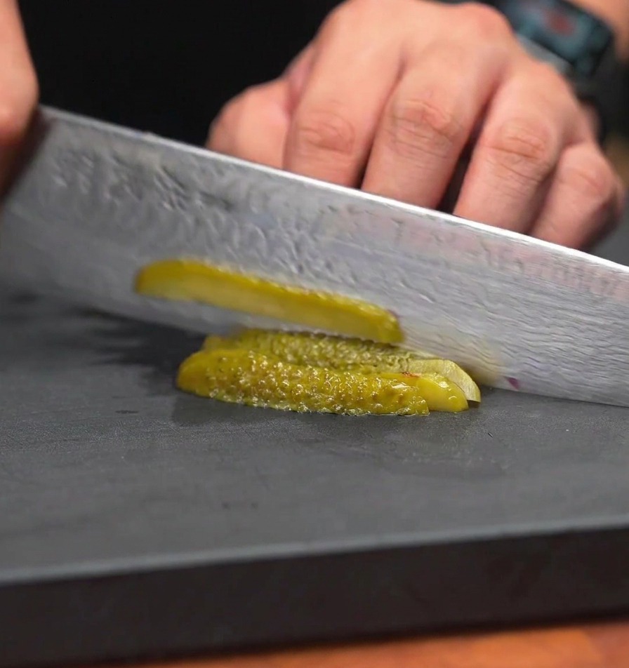 Un couteau de chef coupant des cornichons en très petits morceaux uniformes sur une surface de travail noire.