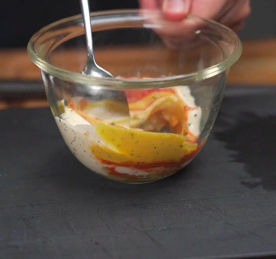 Une cuillère mélangeant du ketchup rouge, de la moutarde jaune et de la mayonnaise blanche dans un bol en verre transparent.