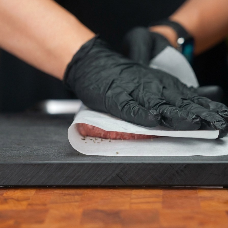 Des mains gantées pressant un steak de bœuf à travers une couche de papier sulfurisé pour laplatir en un disque large et fin.