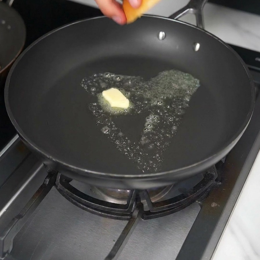 Une main plaçant un pain burger brioché dans une poêle avec un cube de beurre fondant.