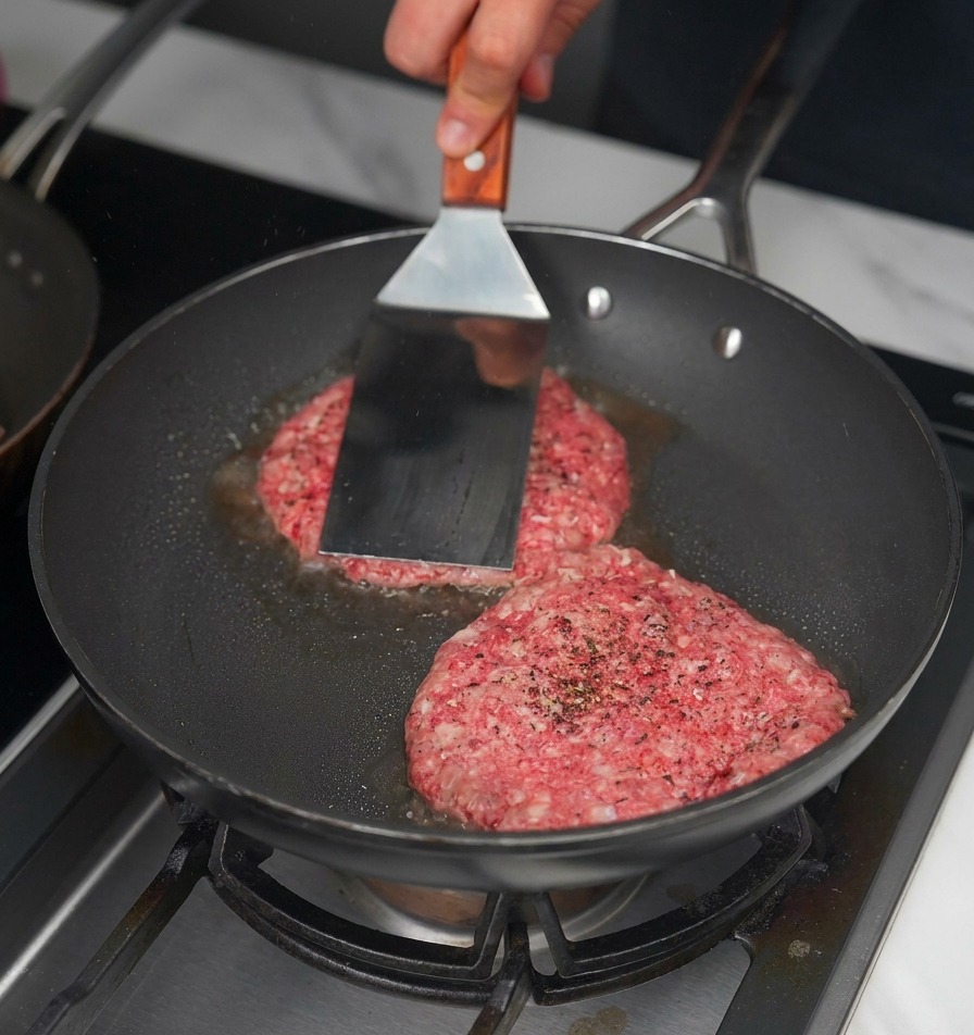 Une spatule métallique pressant deux steaks de bœuf crus dans une poêle noire pour créer une croûte.