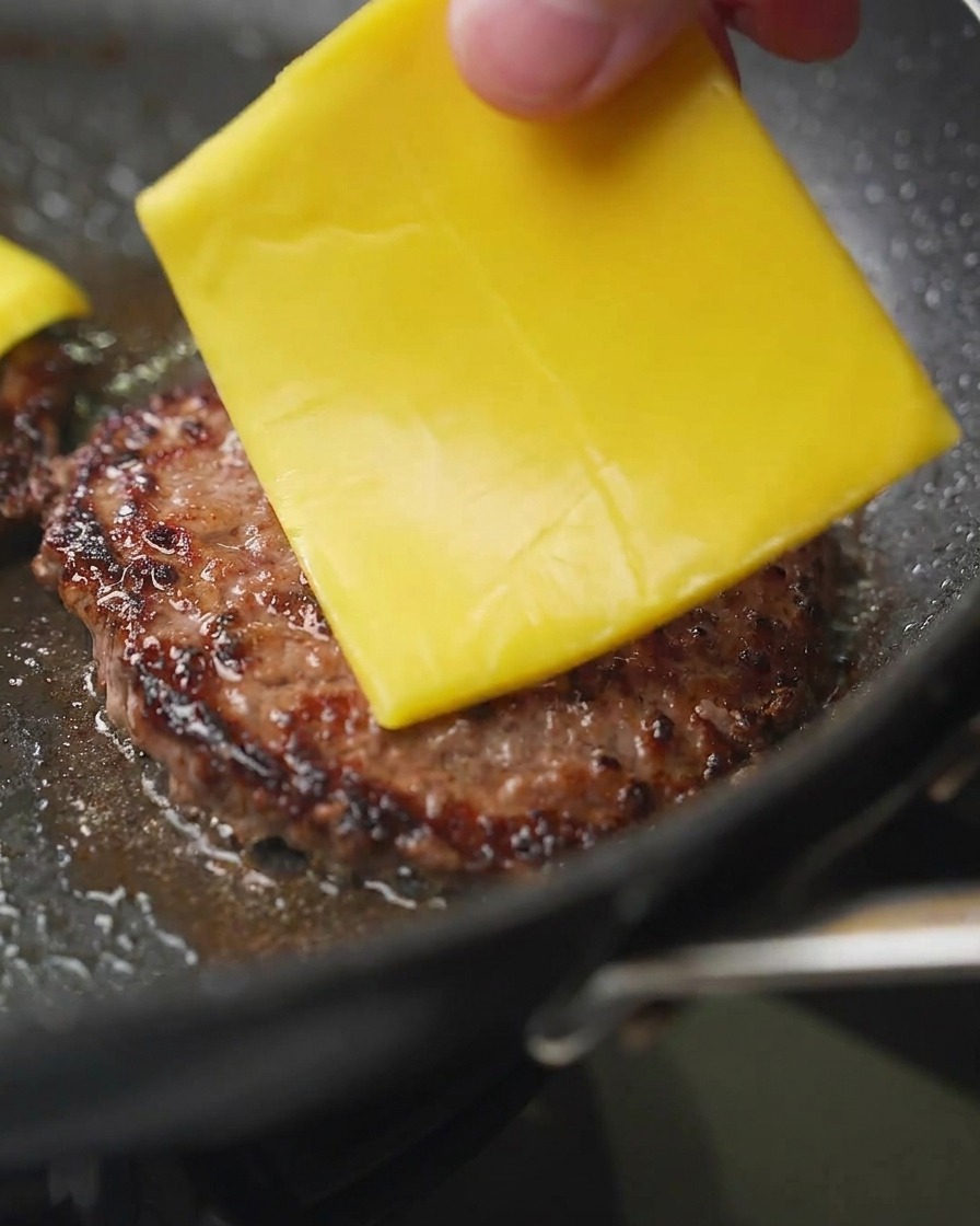 Gros plan dune main déposant une tranche de cheddar jaune sur un steak de bœuf doré dans une poêle.