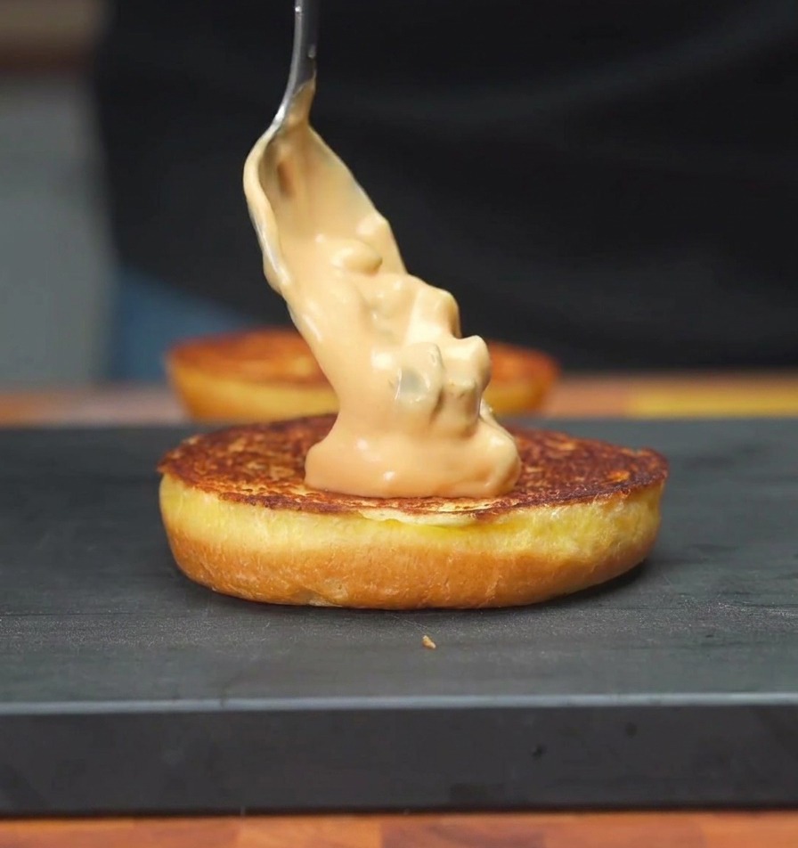 Une cuillère étalant une sauce crémeuse aux cornichons sur la face toastée dun pain burger.