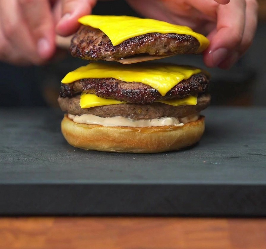 Une main déposant une pile de deux steaks au fromage fondu sur un pain burger nappé de sauce.