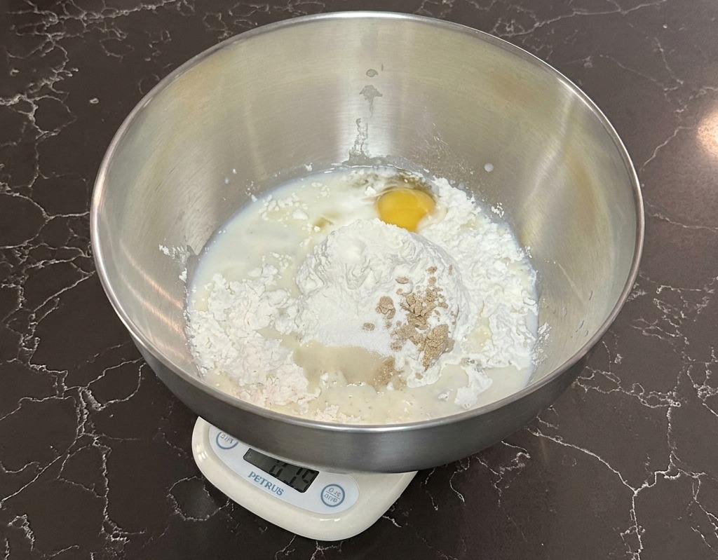 Un bol mélangeur en inox sur une balance digitale contenant de la farine, du lait, un œuf et de la levure.
