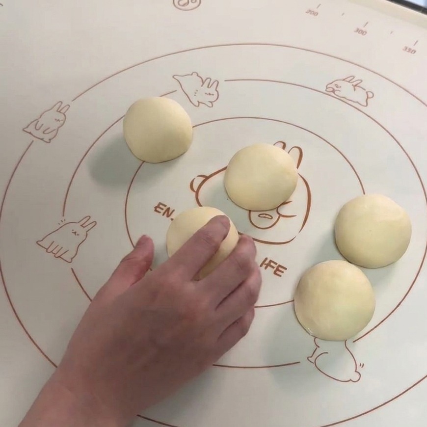Six boules de pâte lisses disposées sur un tapis de cuisson, avec une main en train de façonner lune des portions.