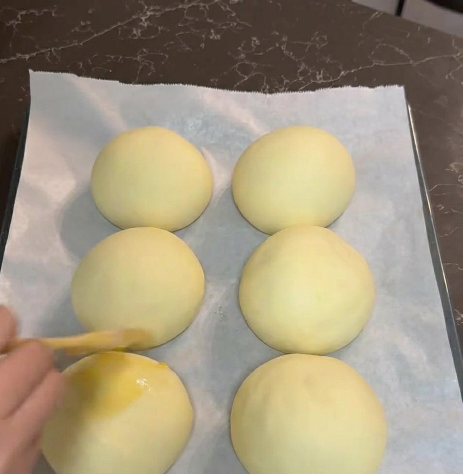 Une main utilisant un pinceau pour badigeonner six boules de pâte levées avec de la dorure à lœuf sur une plaque de cuisson.