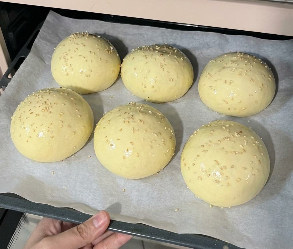 Une plaque de cuisson contenant six pains à burger parsemés de sésame, enfournée dans un four préchauffé.