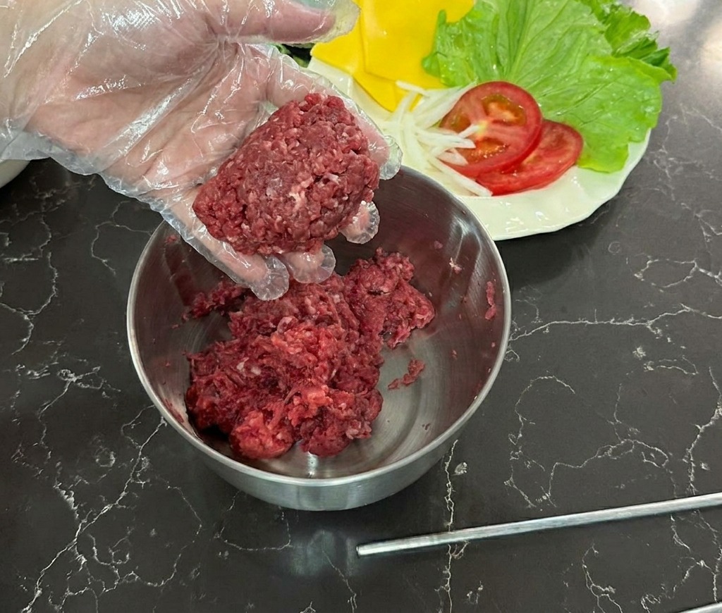 Une main gantée tenant une boule de bœuf haché cru au-dessus dun bol en inox.