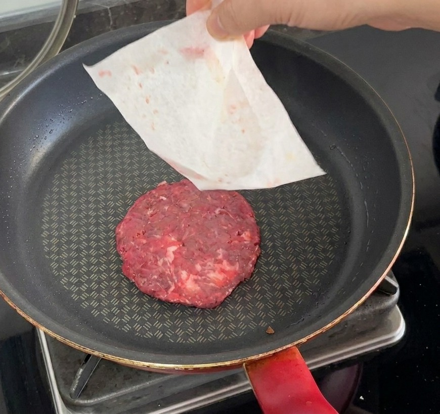 Un steak de bœuf cru déposé dans une poêle chaude, le papier sulfurisé étant retiré manuellement.