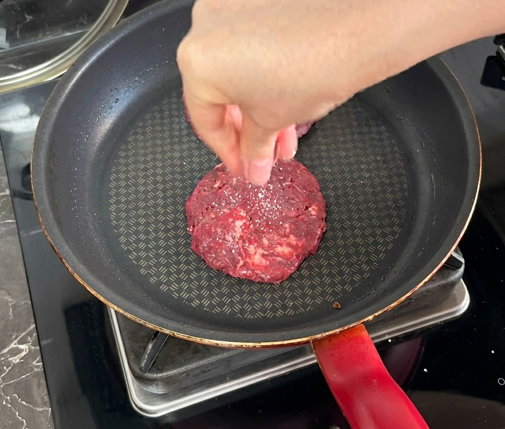 Une main saupoudrant du sel sur un steak de bœuf en train de griller dans une poêle antiadhésive sombre.