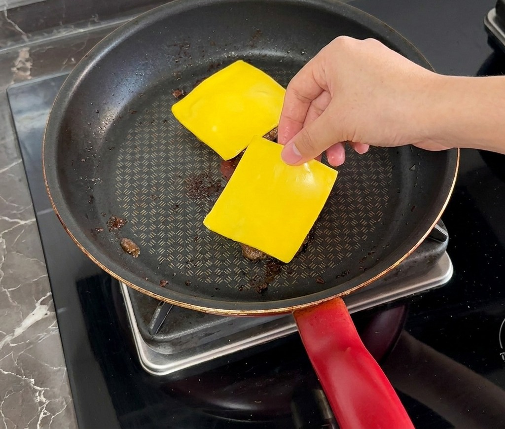 Une main déposant une tranche de fromage jaune sur un steak de bœuf cuit dans une poêle.