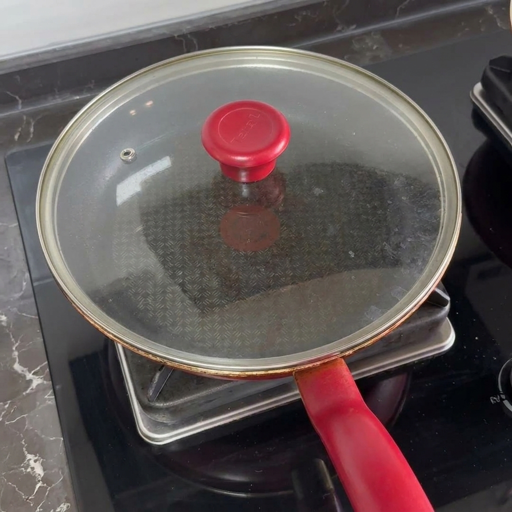 Un couvercle en verre avec une poignée rouge couvrant une poêle pour faire fondre le fromage sur les burgers.