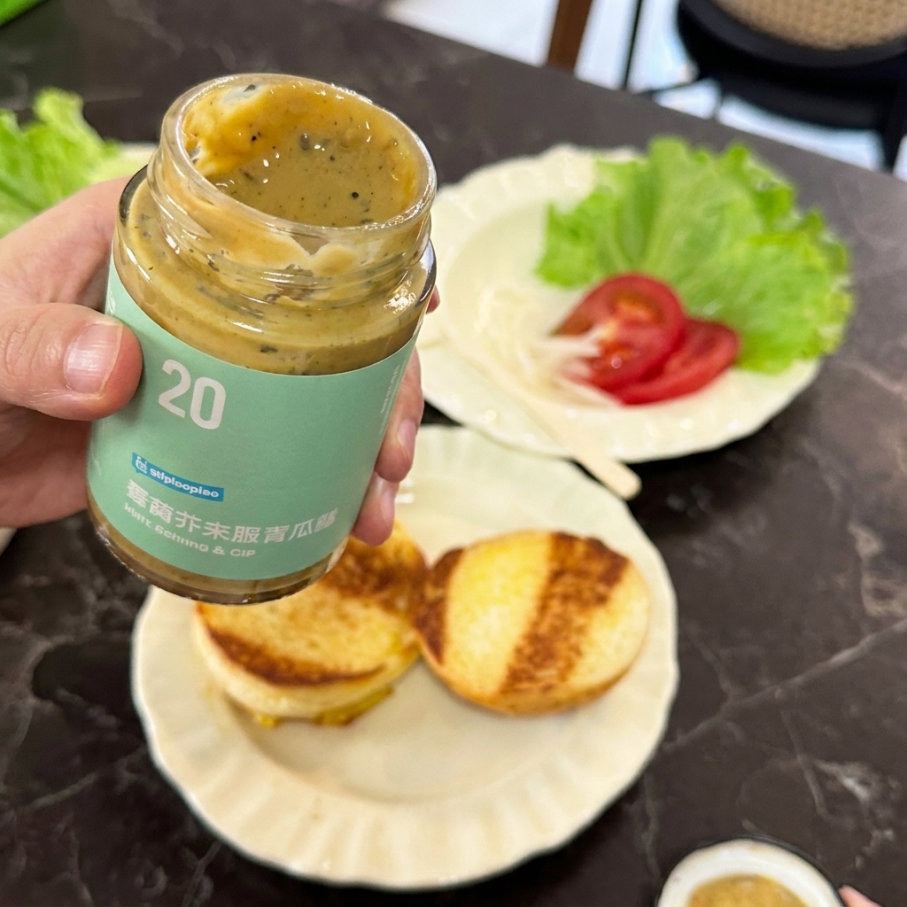 Une main tenant un pot de sauce moutarde et cornichons au-dessus de pains à burger dorés.