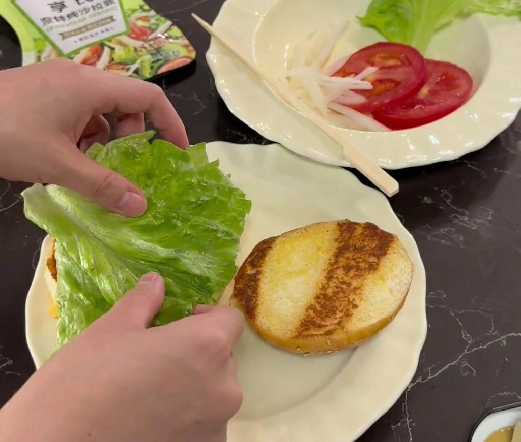 Mains déposant une feuille de laitue fraîche sur un pain à burger toasté posé sur une assiette blanche.