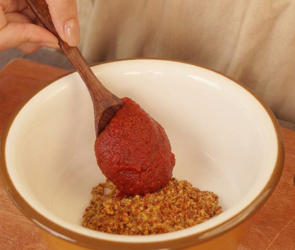 Cuillère en bois ajoutant une sauce pimentée rouge à un bol blanc contenant de la moutarde à lancienne et de la mayonnaise.