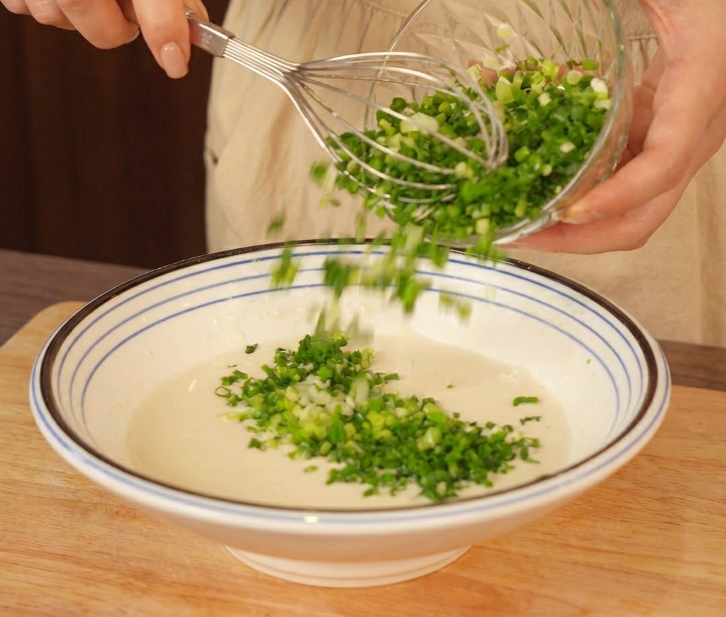 Grand bol de pâte pâle mélangée au fouet alors quune généreuse poignée de ciboule hachée est ajoutée.