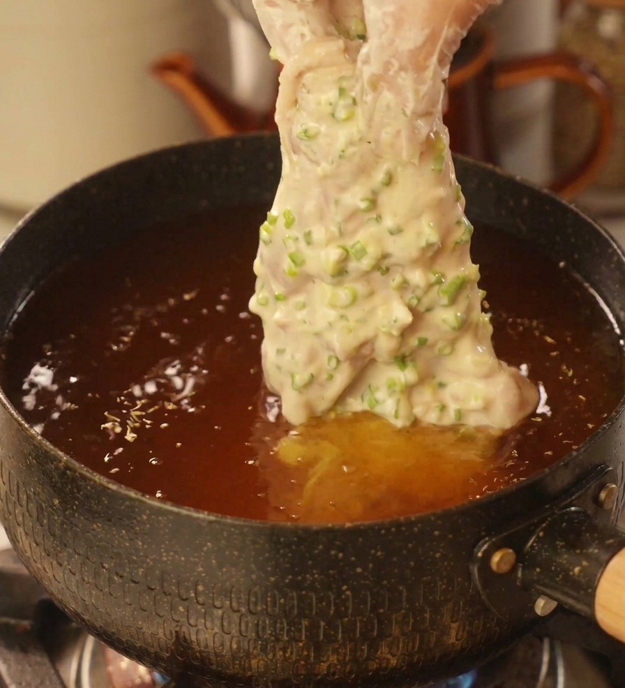 Main avec un gant en plastique déposant un morceau de poulet généreusement enrobé de pâte à la ciboule dans une marmite dhuile chaude.