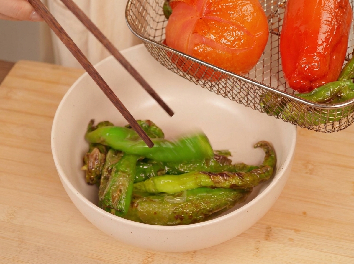 Des baguettes en bois saisissant un piment vert grillé pour le placer dans un bol, avec la tomate carbonisée visible dans le panier de lair fryer.