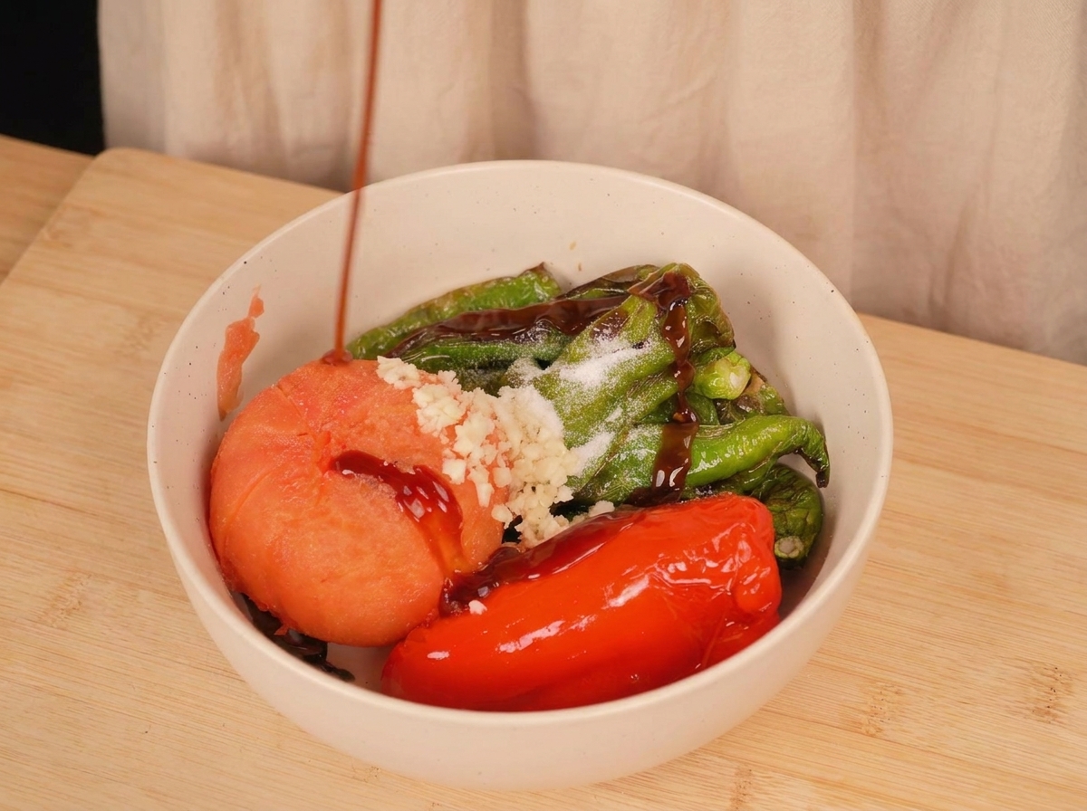 Sauce dhuître sombre versée sur des piments grillés, une tomate, de lail émincé et du sel dans un bol blanc.