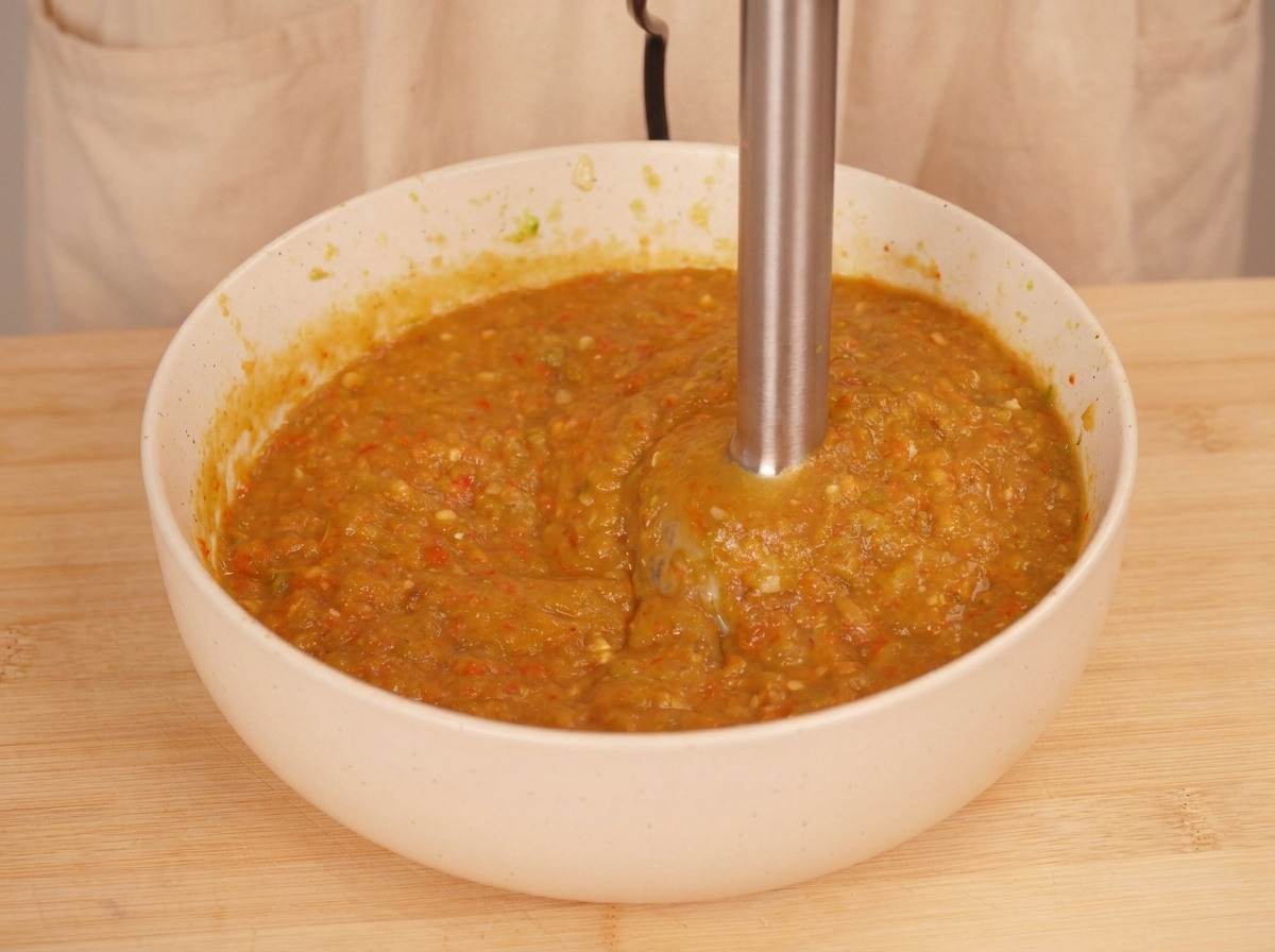 Un mixeur plongeant utilisé dans un bol blanc pour transformer les légumes grillés en une sauce épaisse et texturée de couleur orange-vert.