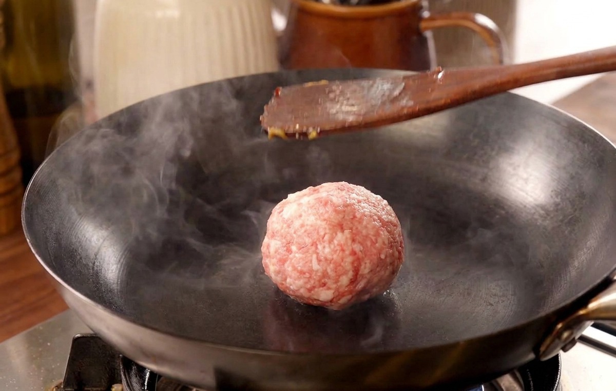 Une spatule en bois pressant une boulette de bœuf dans une poêle brûlante, libérant un nuage de vapeur.