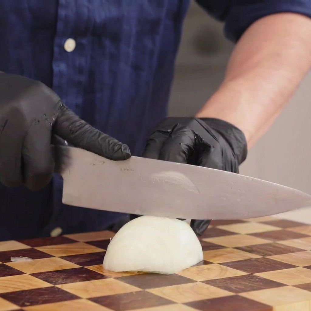 Gros plan dune personne avec des gants coupant finement un oignon blanc sur une planche en bois.