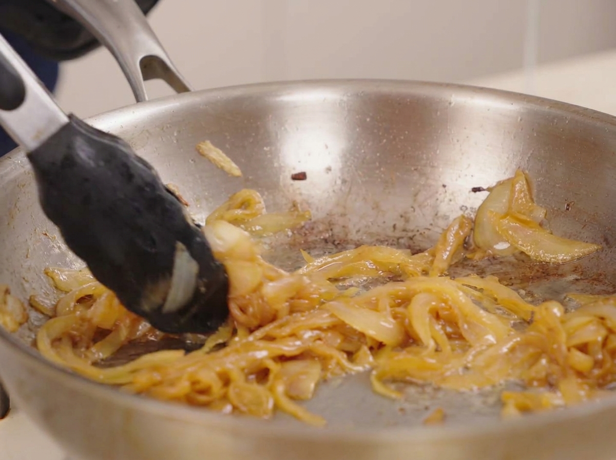 Pinces mélangeant des oignons caramélisés brun doré et fondants dans une poêle scintillante.
