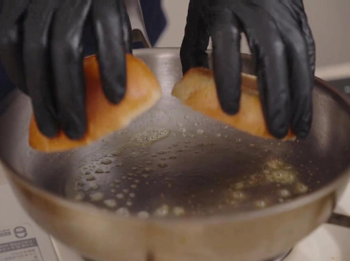 Mains gantées déposant deux moitiés de pain brioché dans une poêle avec du beurre fondu.