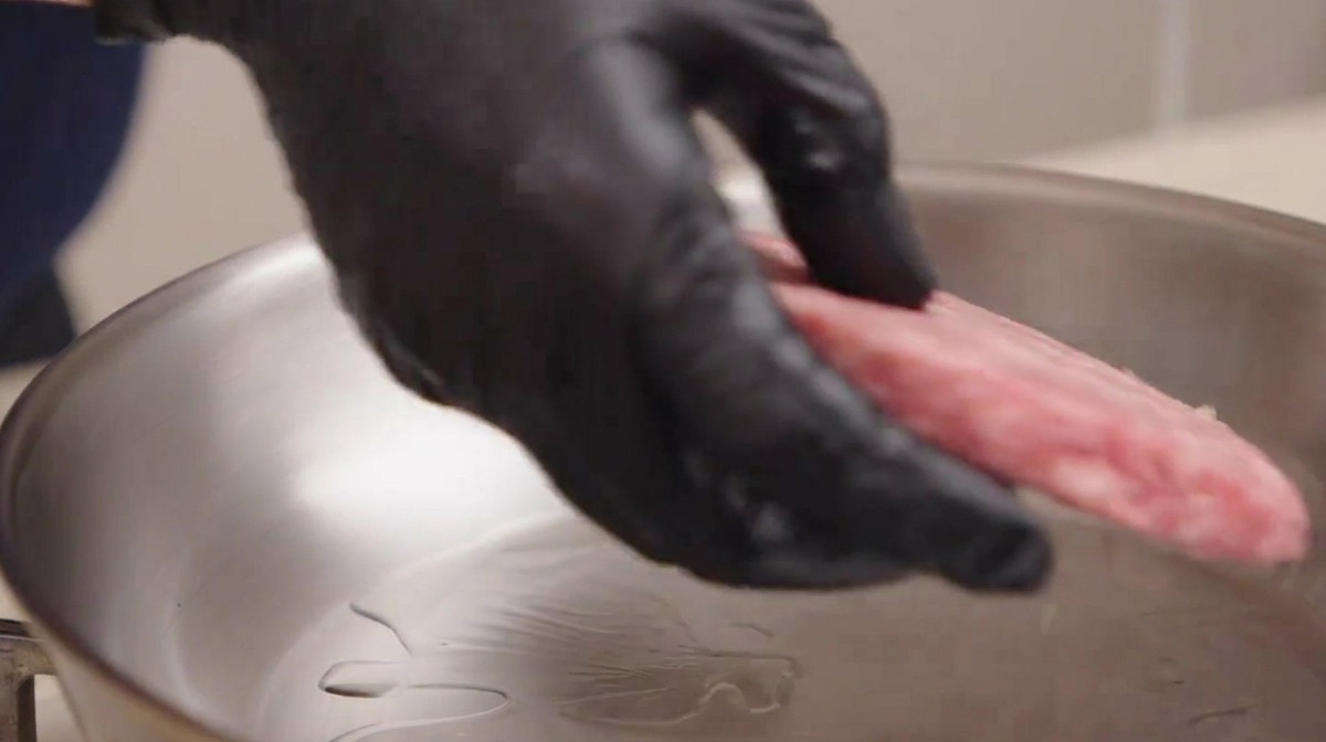 Main gantée déposant un steak de bœuf persillé cru dans une poêle en inox fumante avec de lhuile.