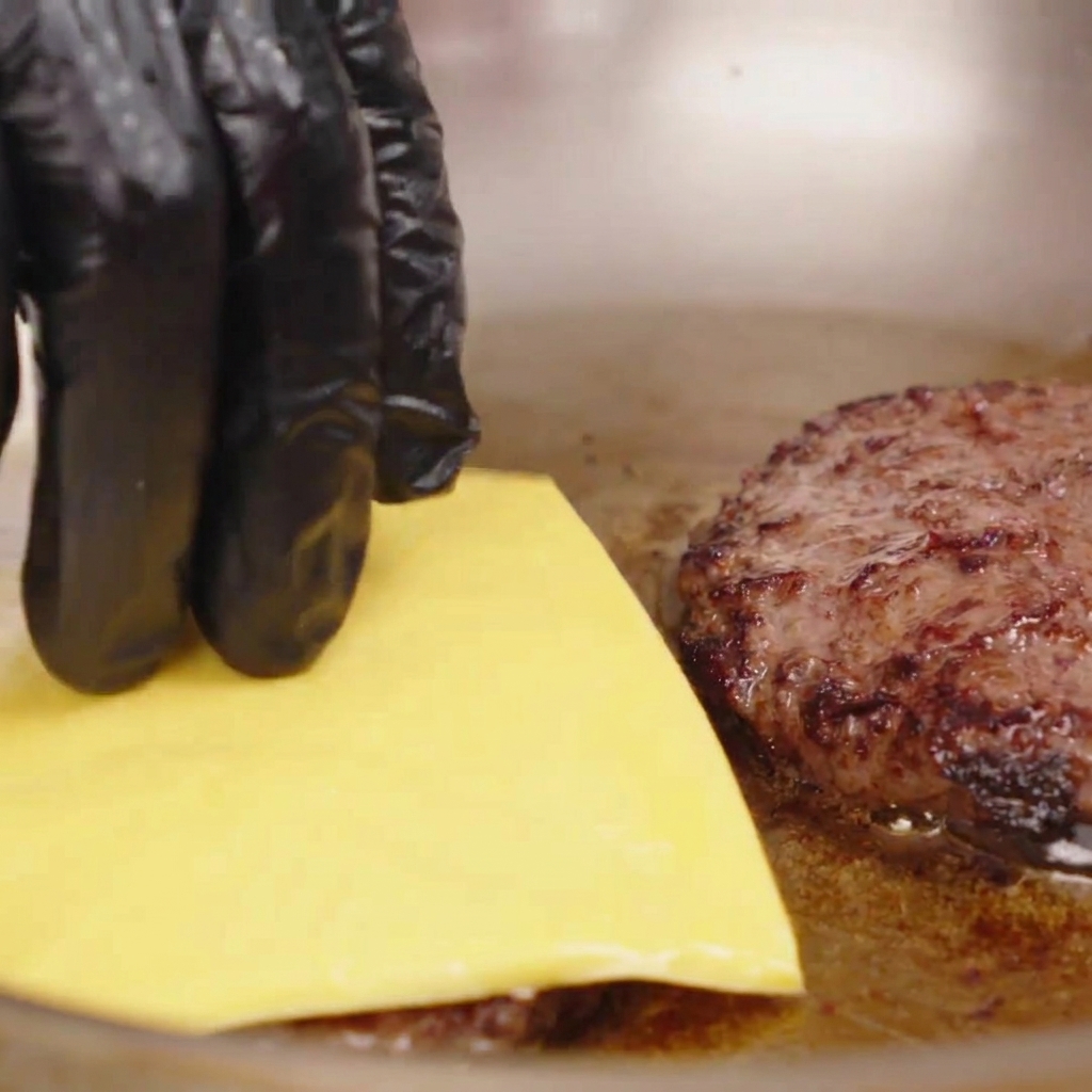 Main gantée déposant une tranche de fromage jaune sur un steak de bœuf chaud dans une poêle.
