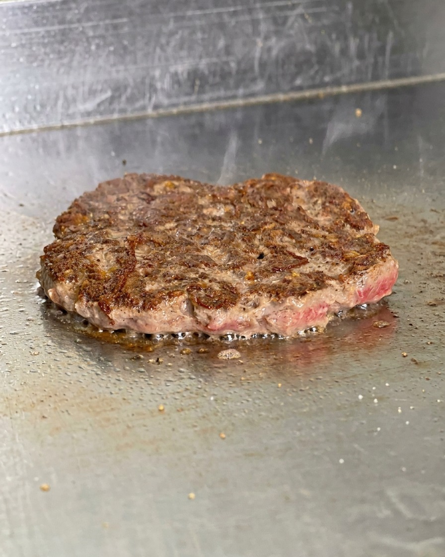 Un steak de burger retourné sur la plaque montrant une croûte brune croustillante avec du gras bouillonnant sur les bords.