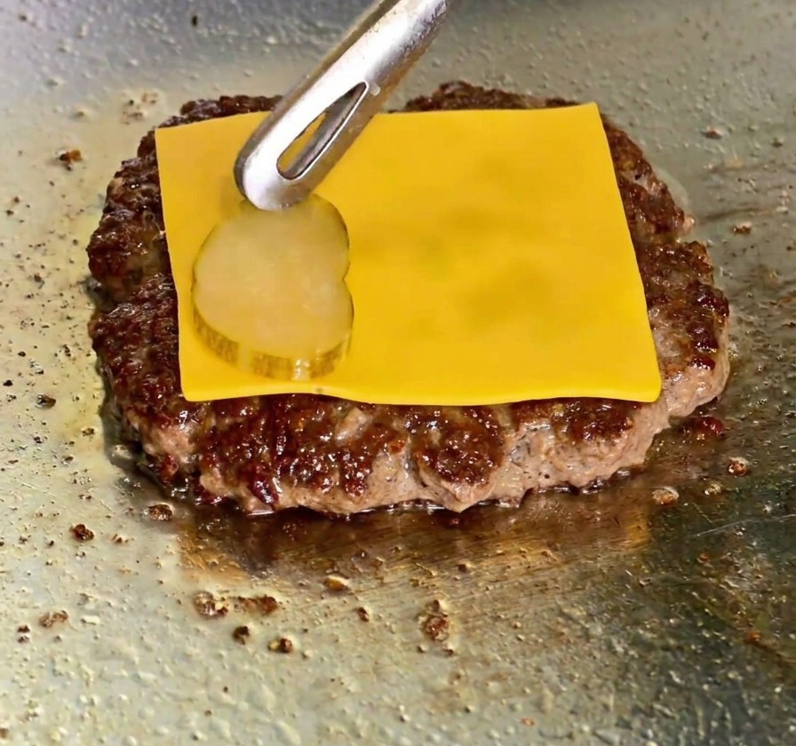 Une tranche de cheddar fondant sur un steak avec des pinces déposant une rondelle de cornichon par-dessus.