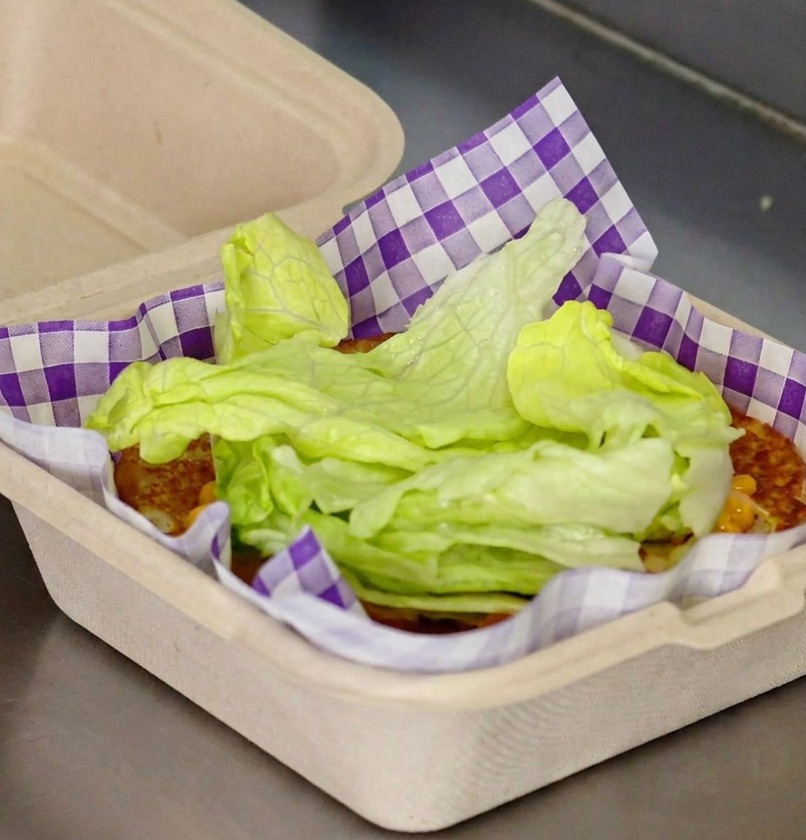 Feuilles de laitue fraîche empilées sur un pain toasté avec de la sauce rouge dans un papier de service à carreaux violets.