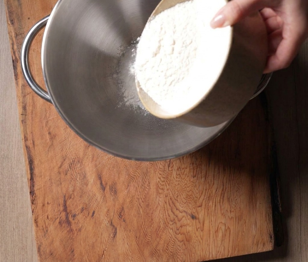 Farine de force blanche versée dun bol en céramique dans la cuve en inox dun robot pâtissier.