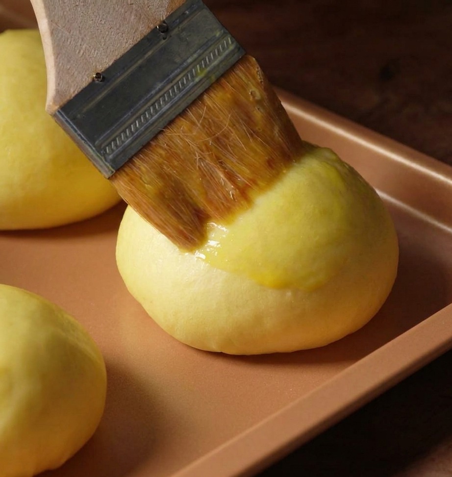 Gros plan dun pinceau appliquant une dorure à lœuf sur un bun lisse avant cuisson.