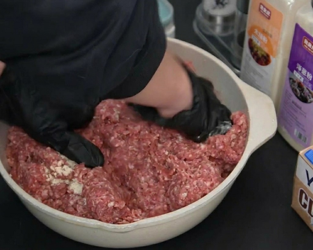 Des mains gantées de noir pétrissant le bœuf haché cru assaisonné dans un plat de couleur claire.