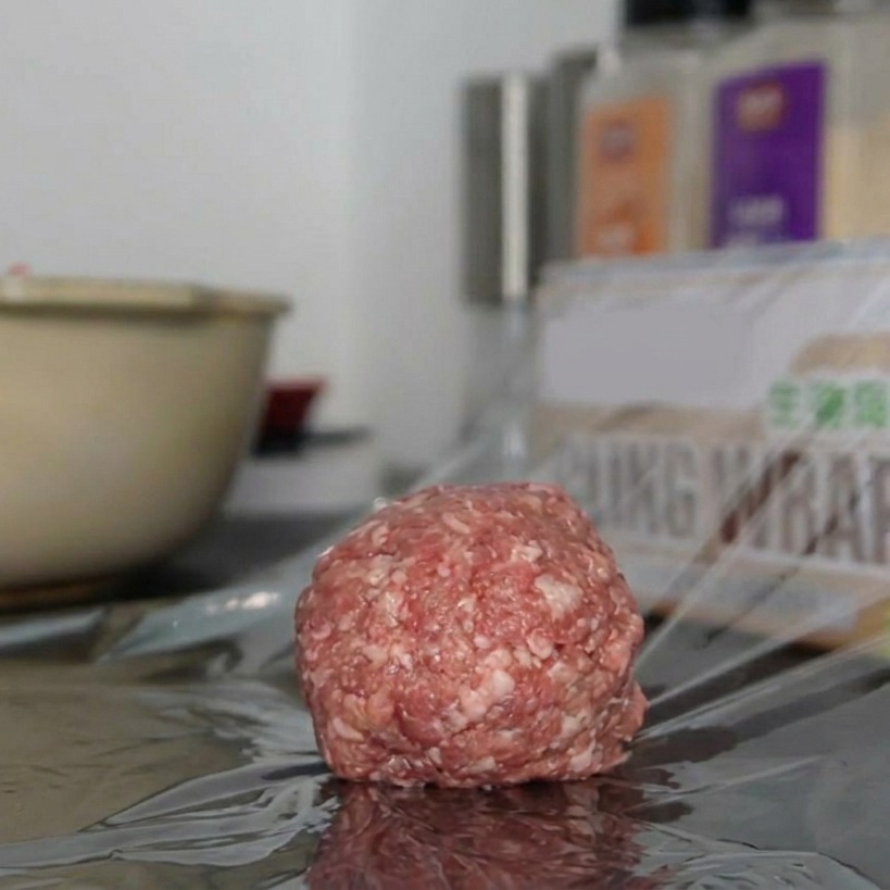 Un beau steak haché cru, parfaitement rond, posé sur un carré de film alimentaire transparent sur le plan de travail.