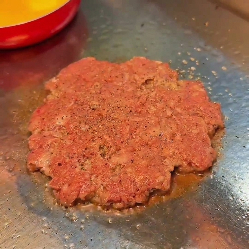 Steak de bœuf smashé en cours de cuisson sur un grill plat, assaisonné de sel et de poivre noir.