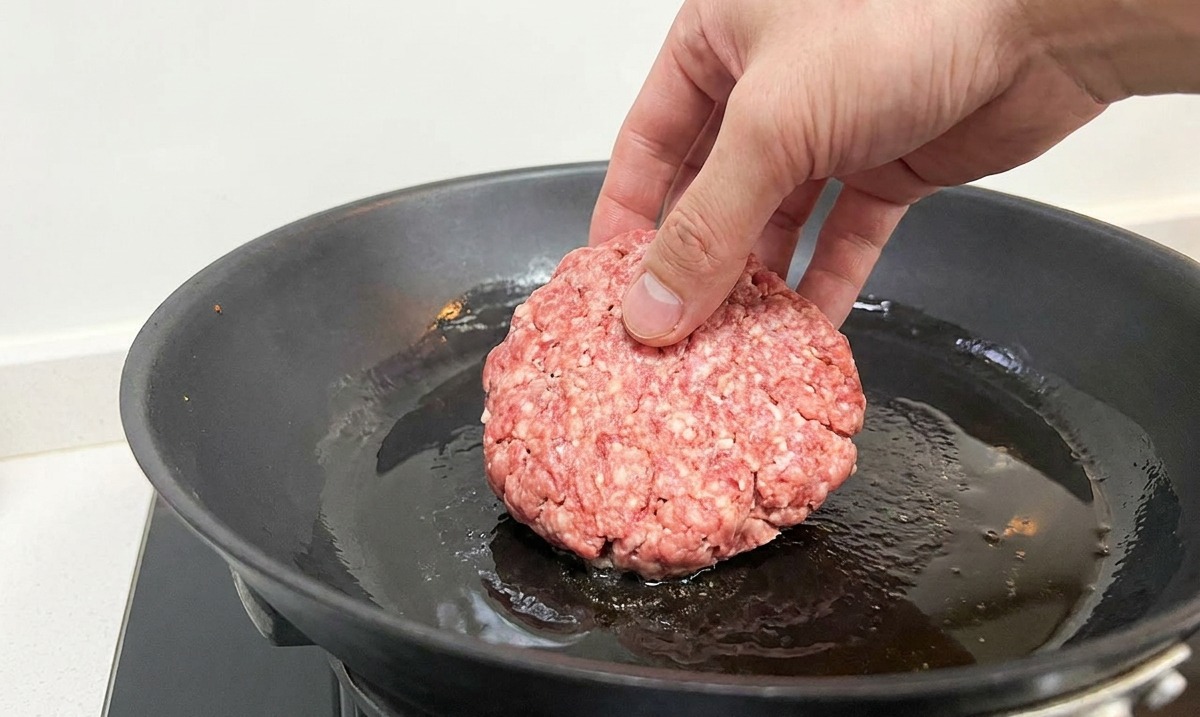 Une main déposant un steak de bœuf cru dans une poêle noire fumante avec un filet dhuile.