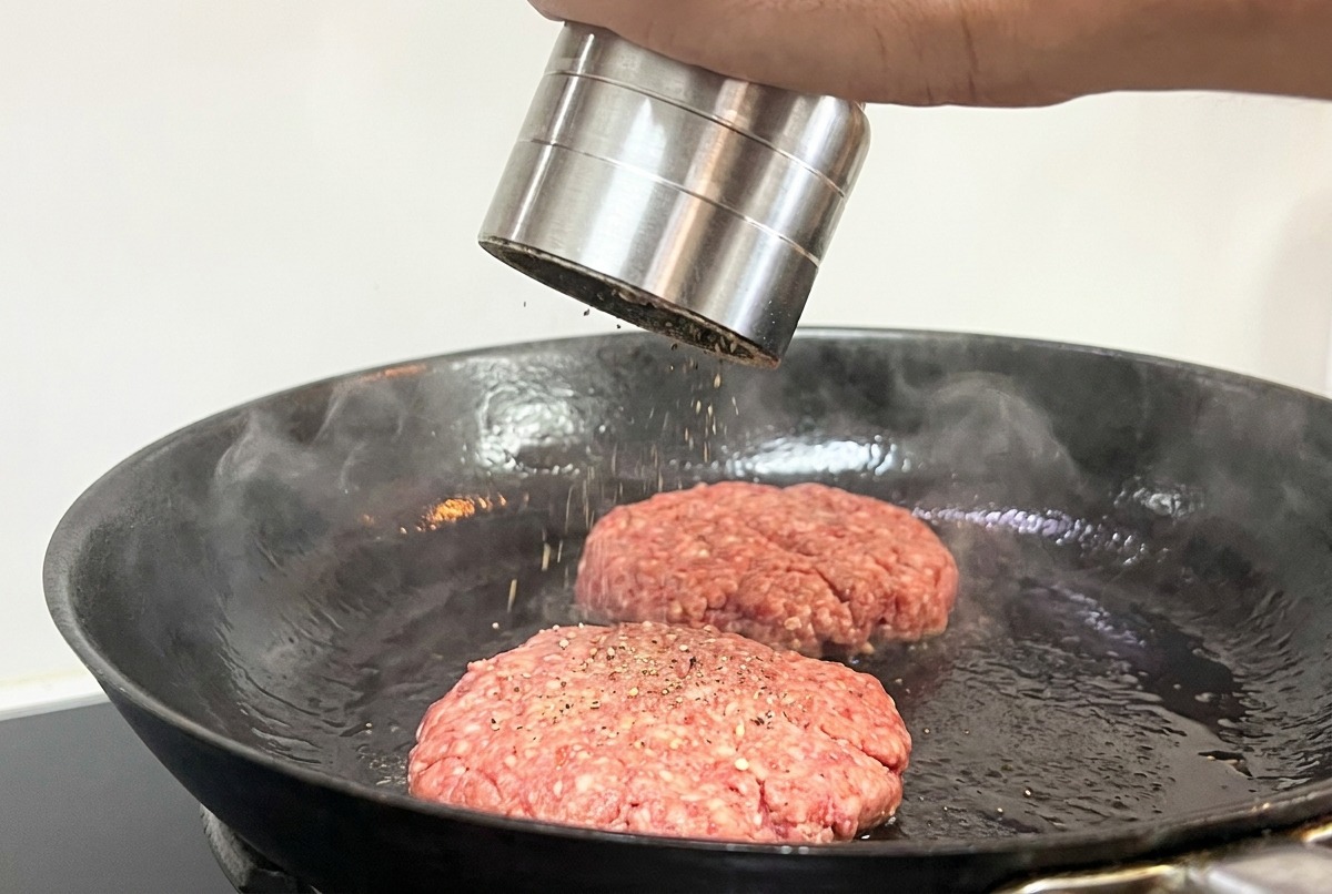 Une main utilisant un moulin à poivre pour assaisonner deux steaks de bœuf en train de cuire.