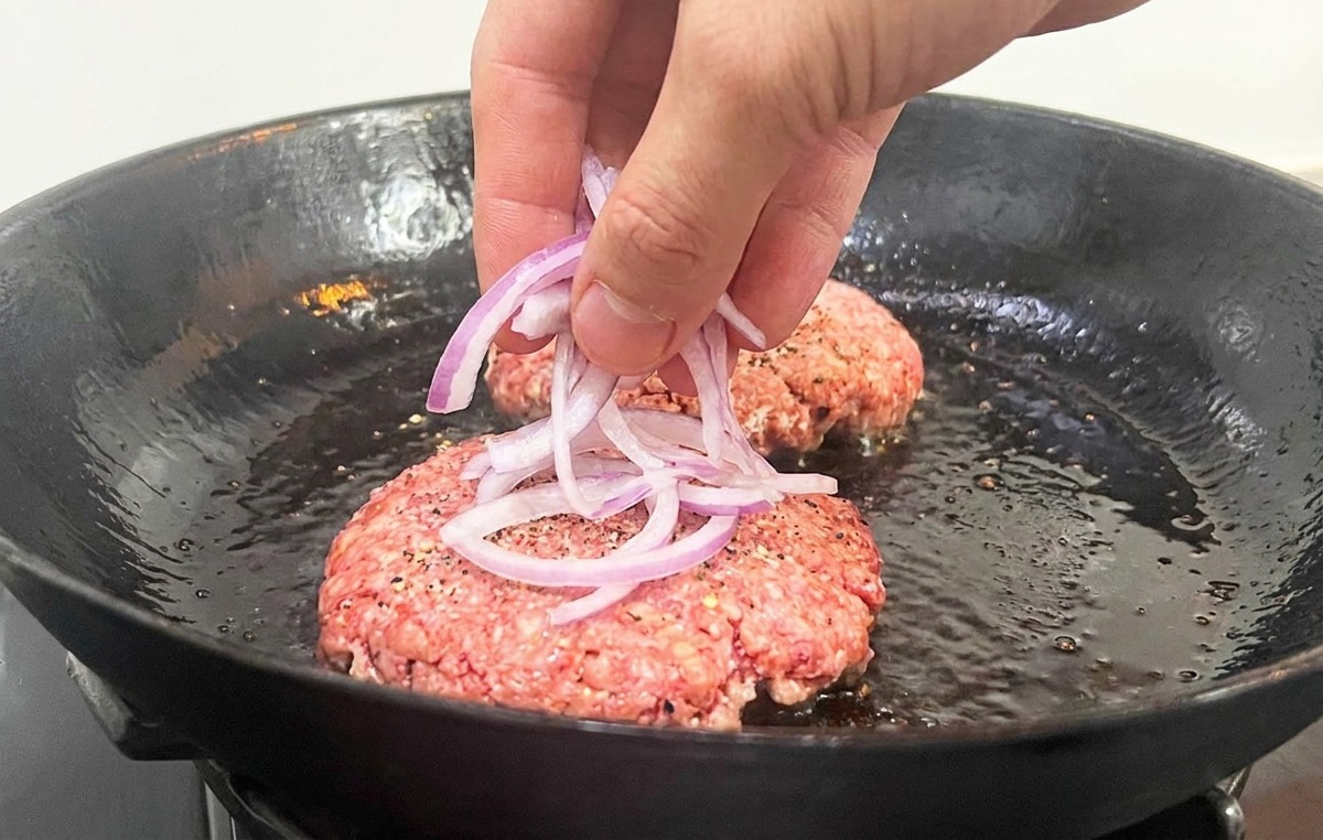 Une main déposant de fines lamelles doignon sur les steaks de bœuf crus dans la poêle.