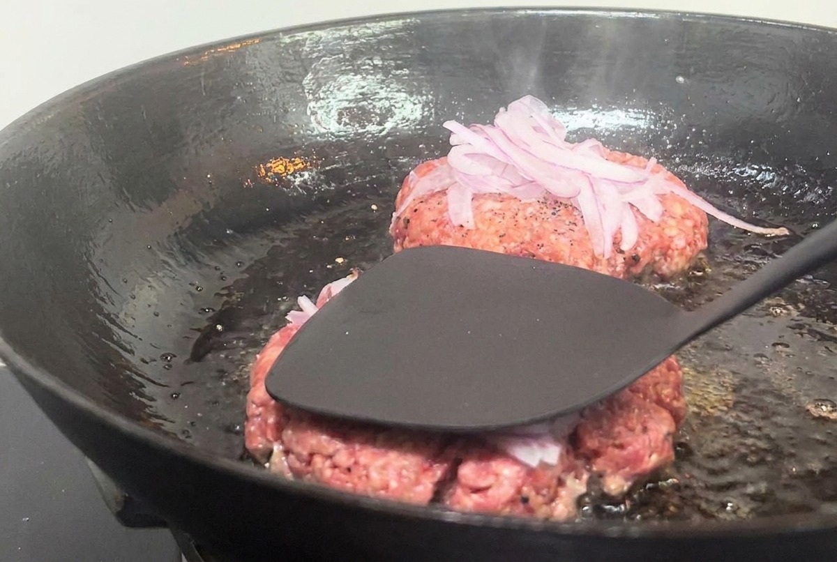 Une spatule métallique pressant fermement un steak de bœuf garni doignons dans une poêle chaude.