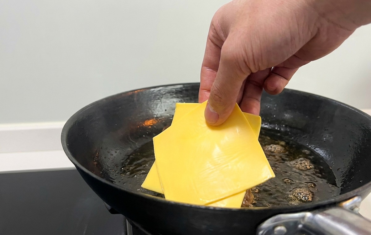 Une main déposant des tranches de fromage jaune sur les steaks saisis dans la poêle.
