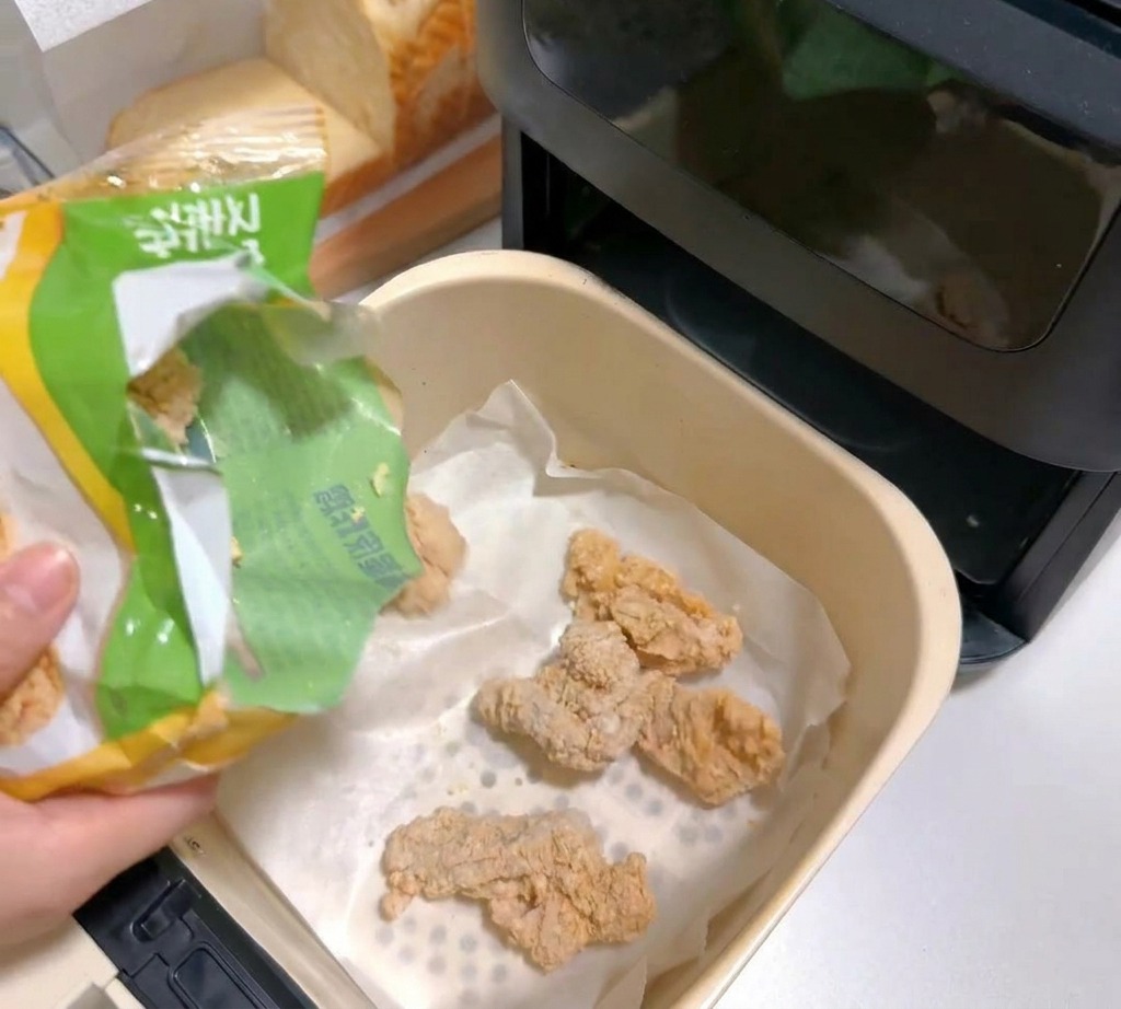Morceaux de poulet pané surgelés versés dans le panier dune friteuse à air tapissé de papier sulfurisé.